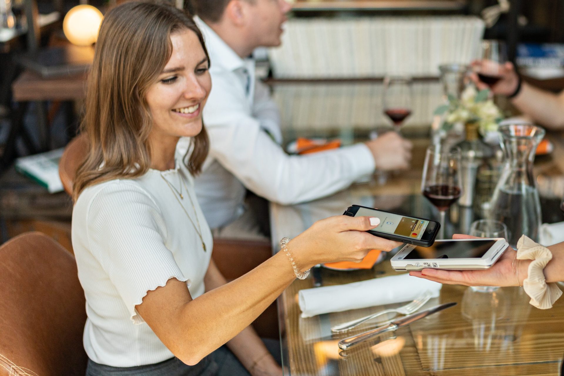 In einem Restaurant sitzen Personen an einem Tisch und trinken gemeinsam Rotwein. Eine Person steht mit einem Bezahlgerät am Tisch und die Kundin bezahlt mit ihrer Kreditkarte auf dem Smartphone, sie lächelt dabei.